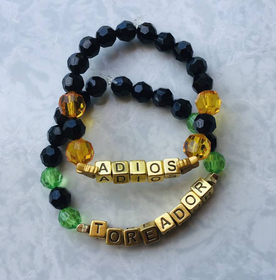 Two simple black kandi singles that say 'ADIOS' and 'TOREADOR'. The 'ADIOS' single has orange beads surrounding the word, while 'TOREADOR' has green.